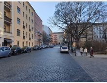 Straßenszene in einem Wohngebiet mit Kopfsteinpflaster, flankiert von mehrstöckigen Altbauten in hellen Farben. Geparkte Autos säumen die Straße, während auf dem Gehweg rechts zwei Fußgänger passieren und kahle Bäume den Bereich säumen.