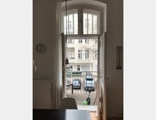 Ausblick von einem hellen Wohnraum mit großen, geöffneten Doppelfenstern in einem weißen Rahmen. Draußen ist ein kleiner Balkon mit Holzboden zu sehen, dahinter geparkte Autos und eine helle Fassade eines gegenüberliegenden Wohnhauses.