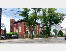 Straßenszene mit Villengebäuden aus verschiedenen Epochen, darunter ein großes rotes Backsteinhaus mit dekorativen Fenstereinfassungen, sowie angrenzenden helleren Häusern. Im Vordergrund befinden sich Bäume, eine gepflasterte Straße mit Straßenbahnschienen und ein hoher Zaun.