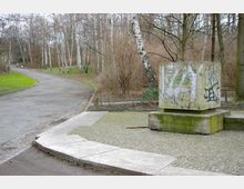 Ein gepflasterter Weg in einem Park mit kahlen Birkenbäumen im Hintergrund. Im Vordergrund ein würfelförmiges, mit Graffiti versehenes Denkmal auf einem steinernen Sockel, umgeben von gepflastertem Boden.