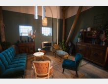 Gemütlich eingerichtetes Wohnzimmer mit grüner Wand, Vintage-Möbeln wie einem blauen Sofa, Sessel und einer Chaiselongue sowie einem runden Beistelltisch. Im Hintergrund sind Holzbalken, ein Kaminschacht und eine Kommode mit dekorativen Elementen und Pflanzen erkennbar.