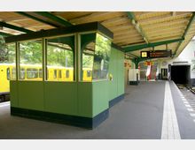 Eine überdachte U-Bahn-Station mit einem grünen Kontrollhäuschen in der Mitte, gelben U-Bahn-Waggons im Hintergrund und Gleisen rechts. Im hinteren Bereich befinden sich ein Treppenaufgang, Hinweisschilder und eine digitale Anzeigetafel mit der Meldung "Hier kein Zugverkehr".