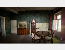 Ein altmodisches Zimmer mit einer grün gemusterten Tapete und abgenutzten Wänden. Im Raum stehen ein runder Holztisch mit vier gepolsterten Stühlen, eine Standuhr, ein holzverzierter Beistelltisch mit einem Landschaftsbild darauf sowie ein geöffnetes Fenster, das Licht hereinlässt.