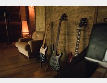 Ein Raum mit Holzwänden und Holzboden, in dem mehrere E-Gitarren und Bässe auf Ständern an der Wand stehen. Links steht ein beigefarbener Sessel neben einer Tischlampe mit warmem Licht und einem Mikrofonständer.