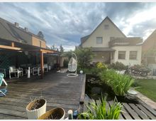 Ein Wohnhaus mit hellem Putz und Satteldach, flankiert von einem gepflegten Garten mit Teich und Holzterrasse. Im Vordergrund stehen Gartenmöbel unter einer überdachten Veranda, und rechts ist ein Motorrad auf einer gepflasterten Fläche zu sehen.