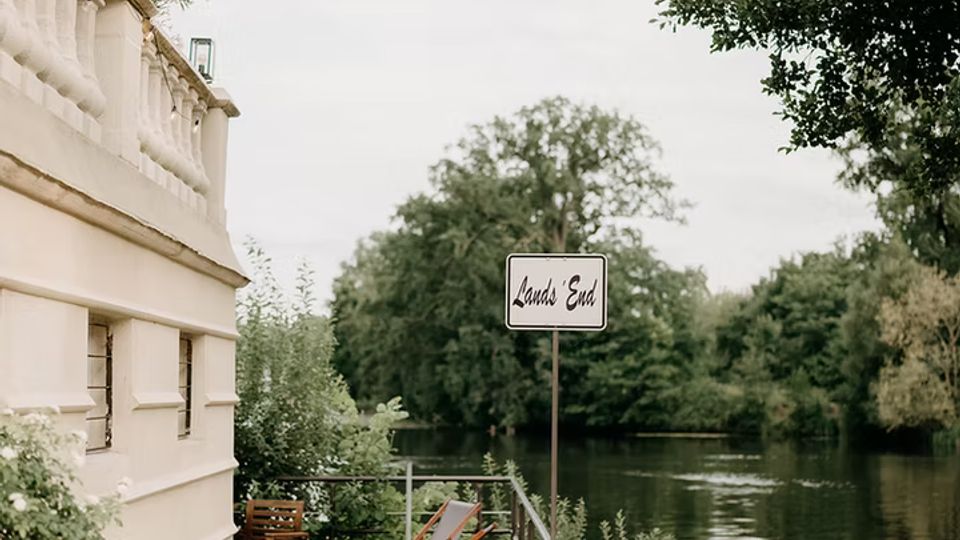 Holzterrasse an einem Flussufer neben einem hellen Gebäude mit Geländer, Liegestühlen und großen Pflanzkübeln. Daneben führt eine Rasenfläche zum ruhigen Wasser, umgeben von Bäumen, mit einem Schild „Land’s End“ am Ufer.