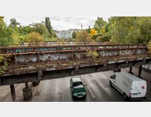 Rostige Eisenbahnbrücke, überwuchert mit Gras und Sträuchern, überspannt eine Straße, auf der ein grünes Auto und ein weißer Lieferwagen fahren. Im Hintergrund sind Bäume, Graffiti und Wohngebäude zu sehen.