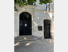 Eingangsbereich eines hellen, historisch wirkenden Gebäudes mit großem Rundbogenportal und schwarzer Glastür, verziertem Stuckrahmen und Steinstufen. Rechts daneben befindet sich eine schmalere schwarze Tür mit der Hausnummer „5“, davor ein gepflasterter Gehweg im Schatten von Baumkronen.