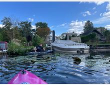 Ein kleiner Wasserkanal mit Seerosen im Vordergrund, ein pinkes Kajak und ein Mann in einem schwarzen Kajak. Im Hintergrund liegt ein weißes Motorboot am Ufer, umgeben von Bäumen, Rasen und Häusern in einer ländlichen Umgebung unter einem blauen Himmel.
