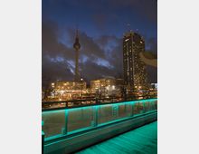 Abendszene in Berlin mit Blick auf den beleuchteten Fernsehturm und das Hochhaus des Park Inn Hotels am Alexanderplatz. Im Vordergrund ein Balkon mit grünlich beleuchteter Glasbrüstung, während im Hintergrund die Stadt bei Nacht mit Straßenlaternen und Gebäuden sichtbar ist.