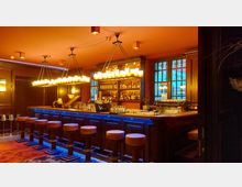 Eine stilvolle Bar mit hölzerner Theke, gepolsterten Barhockern und einer Beleuchtung aus Kerzenleuchtern an der Decke. Im Hintergrund Regale mit Flaschen und ein Fenster, das einen schwachen Blick nach draußen ermöglicht.