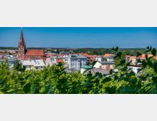 Stadtansicht mit einer Kirche im Zentrum, deren rotes Dach und Glockenturm hervorstechen, umgeben von Wohnhäusern mit weißen und roten Dächern; im Vordergrund grüne Bäume und im Hintergrund ein Wald sowie ein klarer blauer Himmel.