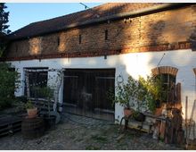 Fassade eines ländlichen Gebäudes mit weiß gestrichener unterer Hälfte und unverputzter Backsteinoberfläche. Vor der Holz-Doppeltür stehen Pflanzen in Töpfen, Holzbalken und andere Materialien; der Boden ist mit Kopfsteinpflaster belegt.