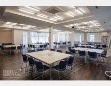 Großer, heller Seminarraum mit mehreren Tischen und dunkelblauen Stühlen in Gruppenanordnung. Der Raum verfügt über dunklen Holzboden, rechteckige Leuchten an der Decke, große Fenster mit weißen Vorhängen und Präsentationselemente wie Flipcharts und Projektor.