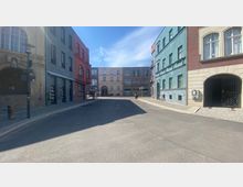 Straßenszene mit mehrstöckigen Gebäuden in verschiedenen Farben, darunter ein rotes Gebäude mit einem "Hotel"-Schild und ein blaues Gebäude mit kleinen Fenstern. Die Straße ist asphaltiert, flankiert von Gehwegen mit Kopfsteinpflaster, und es sind Fahrräder sowie ein Roller zu sehen.