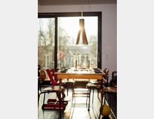 A dining area with a wooden table set for a meal, surrounded by wooden chairs, and illuminated by sunlight streaming through a large glass sliding door. A modern, cone-shaped pendant light hangs above, and the outdoor scenery is partially visible.
