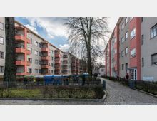 Wohnsiedlung mit mehrstöckigen Wohngebäuden in Beige und Rosa, umgeben von Gehwegen aus Kopfsteinpflaster, kleinen Grünflächen und Zäunen. Mittig stehen Bäume sowie blaue Müllcontainer auf einem abgesperrten Bereich.