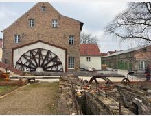 Backsteinbauwerk mit einem großen, dekorativen Metallrad an der Fassade, daneben ein kleineres weißes Gebäude mit rotem Ziegeldach. Im Vordergrund sind historische Mauerreste und alte Zahnräder zu sehen, im Hintergrund weitere Backsteingebäude mit grünen Toren.