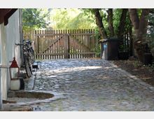 Pflastersteingasse mit links an einer Hauswand abgestellten Fahrrädern und einer roten Laterne; im Hintergrund ein Holztor sowie Bäume und rechts Mülltonnen an einem schattigen Bereich.