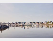 Eine Reihe von kleinen Häuschen mit spitzen Dächern, die in verschiedenen Farben gestrichen sind, stehen entlang eines Hafens; im Vordergrund liegt ein ruhiges Gewässer mit festgemachten Booten, und Stege führen von den Häusern ins Wasser.