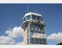 Ein Kontrollturm mit verglasten Fenstern und einer Aussichtsplattform, beschriftet mit den Buchstaben "EDAZ", unter klarem blauen Himmel mit vereinzelten Wolken.