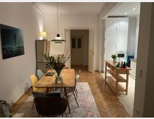 Essbereich in einer Wohnung mit Holztisch, umgeben von farblich unterschiedlichen Stühlen, auf einem Teppich mit Muster. Im Hintergrund eine offene, weiße Küche mit hoher Schrankwand und hölzerner Kücheninsel; Stuckverzierungen an der Decke betonen den Altbaucharakter.