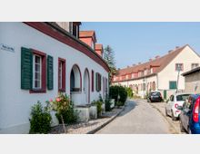 Eine gepflasterte Straße in einer Wohnsiedlung mit Reihenhäusern im traditionellen Stil; die Häuser haben weiße Fassaden, rote Ziegeldächer, grüne Fensterläden und Rundbögen an den Eingängen. Am Straßenrand parken mehrere Autos, und vor den Häusern wachsen Rosen und Büsche.