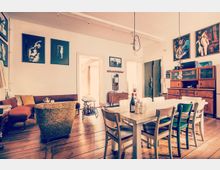 Wohnzimmer mit Vintage-Einrichtung, bestehend aus einer weißen Holztafel mit Stühlen, einer Eckcouch, einem Sessel und einem Servierwagen. An den weiß gestrichenen Wänden hängen mehrere Kunstwerke, und ein großer Holzschrank sowie Holzboden vervollständigen den Raum.