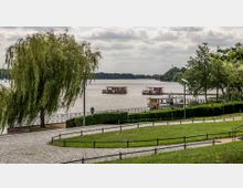 Parklandschaft mit gepflastertem Weg, einem großen Baum und Blick auf einen See mit schwimmenden Holzplattformen und einem kleinen Boot am Ufer. Im Hintergrund sind bewaldete Ufer und wolkiger Himmel zu sehen.