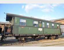 Historischer grüner Eisenbahnwaggon mit mehreren Fenstern, dekoriert mit weißen Gardinen. Der Waggon steht auf Gleisen vor einem Backsteingebäude, vermutlich einem Bahnbetriebswerk.