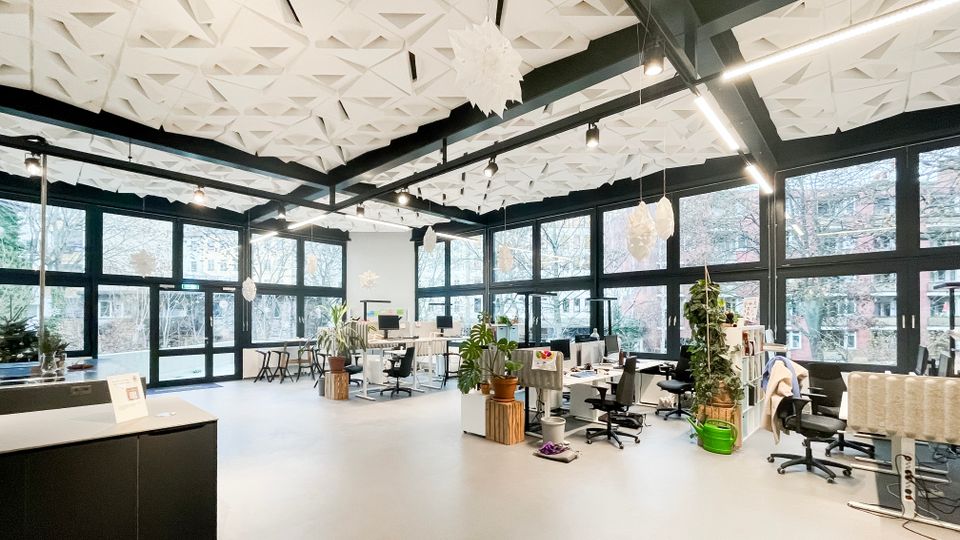 Helles, offenes Großraumbüro mit bodentiefen Fensterfronten und schwarzem Stahlträger-Rahmen, das viel Tageslicht hereinlässt. Auffällig sind die geometrische, weiße Deckenverkleidung, lineare Hängeleuchten, mehrere Arbeitsplätze mit Rollstühlen sowie Pflanzen und Regale als Raumteiler.