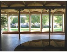 Innenansicht eines Gebäudes mit einer gewölbten Ziegeldecke und großen bodentiefen Fenstern, die einen Blick auf eine grüne Gartenlandschaft mit Bäumen und blühenden Sträuchern bieten. Im Vordergrund sind Metallstützen und ein dunkles, reflektierendes Element sichtbar.