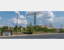 Straßenszene mit einer Bushaltestelle am Straßenrand, einem hohen Strommast mit Überlandleitungen und einem Industriegelände im Hintergrund, inklusive eines großen Backsteingebäudes mit mehreren Industrieschornsteinen. Die Umgebung ist von Bäumen und Sträuchern umgeben, und der Himmel ist leicht bewölkt.