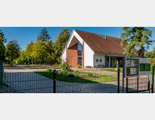 Modernes Gebäude mit rotem Ziegeldach und einer Kombination aus weißer und roter Fassade, umgeben von Grünflächen mit vereinzelten Pflanzen. Im Vordergrund ist ein Zaun sowie eine Infotafel sichtbar, während im Hintergrund ein parkähnliches Areal mit Bäumen zu erkennen ist.