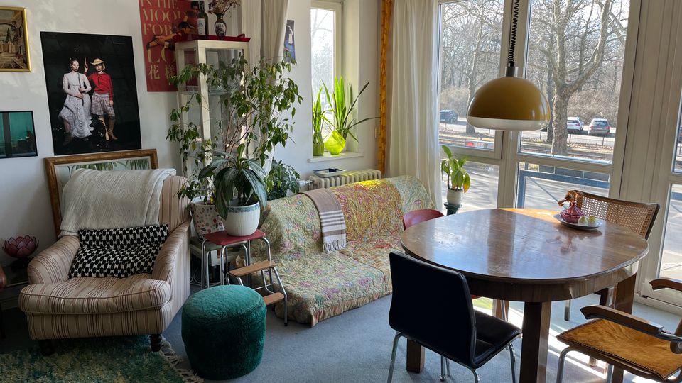 Bright living and dining area with large corner windows and sheer curtains, overlooking trees and a street outside. The room has a round wooden dining table under a dome pendant light, a patterned sofa and armchair, indoor plants, and framed artwork on white walls.