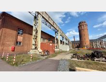 Ehemaliges Industrieareal mit Backsteingebäuden, darunter ein hoher runder Turm mit Fensterreihen. Im Vordergrund stehen Container, Industriegeräte und eine verrostete Metallkonstruktion, die entlang des Geländes verläuft.