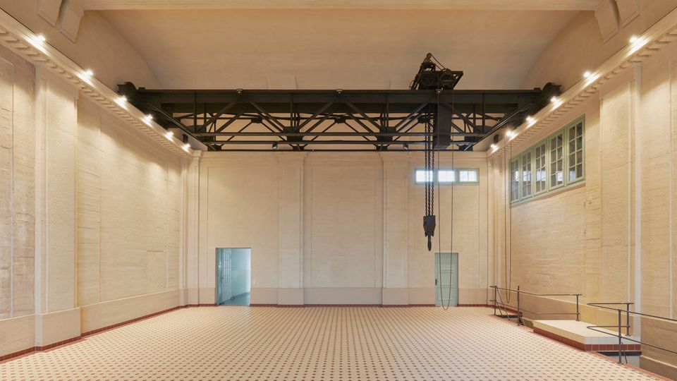 Großer, heller Innenraum mit hohen Decken, cremefarbenen Wänden und einem schwarz lackierten Trägergerüst mit einem Kettenzug. Der Boden besteht aus einem karoförmigen Fliesenmuster, und seitlich befinden sich kleine Fenster sowie Türen, die in andere Räume führen.