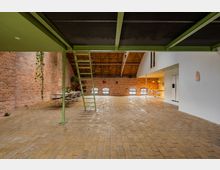 Großer loftartiger Raum mit Ziegelsteinwänden, gelbem Fliesenboden und grünen Metallstützen. Im Hintergrund befindet sich ein Esstisch mit Stühlen, eine weiße Wand trennt eine offene Küche, und eine grüne Metalltreppe führt zu einer Zwischendecke.