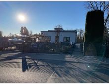 Modernes Wohnhaus mit flachem Dach und einer weißen Fassade, umgeben von einem Metallzaun. Im Vordergrund befindet sich eine Einfahrt, daneben hohe Sträucher und ein gepflegter Garten; die Sonne steht tief am klaren Himmel.