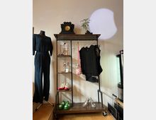 Ein offener Kleiderständer aus Metall mit Holzregalen, darauf verschiedene Accessoires wie eine alte Uhr, eine Pflanze und Schmuck. Darunter hängen schwarze Kleidungsstücke, und auf den unteren Regalböden stehen bunte Schuhe. Links daneben steht eine schwarze Schneiderpuppe mit einem dunkelblauen Kleid.