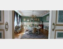 Blick durch eine geöffnete Flügeltür in ein historisch eingerichtetes Salon- oder Speisezimmer mit grüner Tapete, weißer Wandvertäfelung und Stuckdecke samt Kronleuchter. In der Mitte steht ein runder Tisch auf einem gemusterten Teppich, umgeben von Polsterstühlen; an den Wänden hängen gerahmte Porträts, daneben stehen eine Vitrine und ein Sofa vor hohen Fenstern mit dunklen Vorhängen.