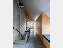 Flur in einem modernen Innenraum mit Beton- und Holzelementen; links eine Betontreppe mit schwarzem Metallgeländer, darüber hängende schlichte Lampen, rechts eine Holzwand mit Schränken. Am Ende des Flurs befindet sich ein Fenster mit Blick ins Freie.