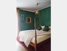 Schlafzimmer mit grün gestrichenen Wänden, einem Holz- Himmelbett mit weißer Bettwäsche und dunkelgrünen Kissen. An der Wand hängen ein vergoldeter Bilderrahmen und ein kleines Regal, darüber eine Hängelampe mit Fransen. Ein roter Teppich bedeckt den Holzboden.
