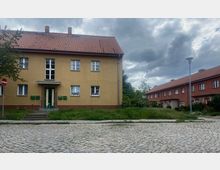 Zweistöckiges Wohngebäude in hellgelber Farbe mit rotem Satteldach und weißen Fenstern, umgeben von einer Grünfläche und Pflastersteinen auf der Straße. Im Hintergrund sind weitere Reihenhäuser in roter Farbe sowie ein bewölkter Himmel sichtbar.