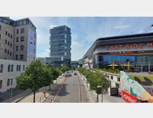 Straßenansicht in einem modernen Stadtteil mit einem hohen Bürogebäude auf der linken Seite und dem geschwungenen Einkaufszentrum "East Side Mall" auf der rechten Seite. In der Mitte verläuft eine von Bäumen gesäumte Straße mit wenigen Fahrzeugen, flankiert von Gehwegen.