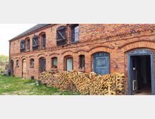 Backsteinbau aus rotem Ziegelstein mit zwei Etagen, arkadenförmigen Türen und Fenstern. Vor dem Gebäude liegt gestapeltes Kaminholz, umgeben von einer grasbewachsenen Fläche.
