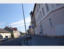 Straßenszene mit mehrstöckigen Wohnhäusern, teils in hellen Farben verputzt, entlang eines Gehwegs auf der rechten Seite. Links sind niedrigere Gebäude mit Ziegeldächern und geparkte Autos sichtbar, ein Straßenschild mit der Aufschrift „Schleusenstraße“ ragt in die Szene.