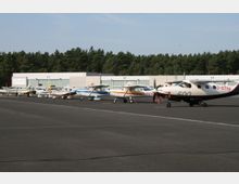 Reihe von Kleinflugzeugen, die auf dem Vorfeld eines Flugplatzes abgestellt sind, vor mehreren Hangars mit der Beschriftung „C1“, „C2“ und „C3“ sowie einem Wald im Hintergrund.