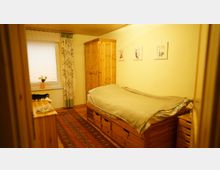 Kleines Schlafzimmer mit Holzmöbeln, darunter ein Einzelbett mit Schubladen darunter, ein Kleiderschrank und eine Kommode. Ein Fenster mit Blumenvorhängen, ein Teppich und helle Wände ergänzen die Einrichtung.