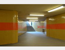 Unterführung mit gelben Kacheln an den Wänden, die von einem roten Streifen durchzogen sind, und Deckenbeleuchtung. Im Hintergrund befindet sich eine Treppe, die ins Freie führt.